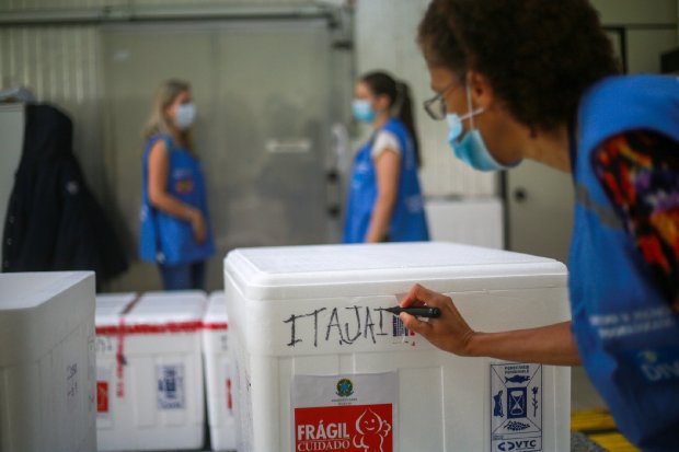 Servidora da saúde escreve Itajaí com marcador preto em caixa térmica branca rotulada como frágil. Ao fundo, duas outras profissionais com coletes azuis e máscaras conversam em ambiente interno, possivelmente um centro de distribuição de vacinas.