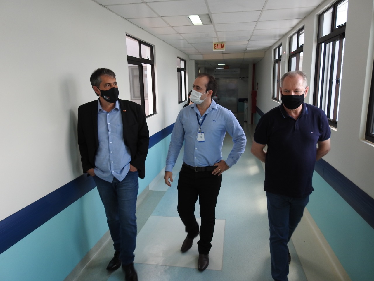 Três homens usando máscaras caminham por corredor de unidade hospitalar; dois deles usam camisa social e o terceiro veste camiseta polo escura. Ambiente iluminado por luz natural vinda das janelas.