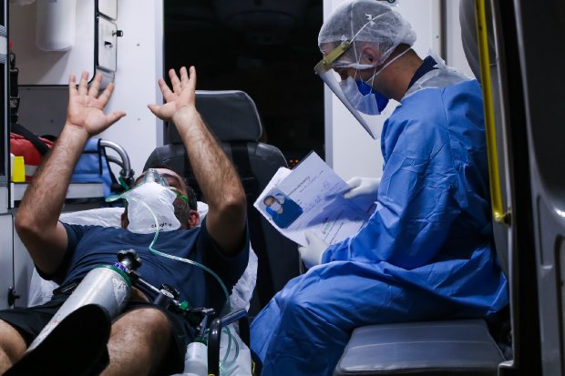Profissional de saúde com equipamento de proteção individual completo atende paciente deitado em maca dentro de uma ambulância. O paciente está com máscara de oxigênio e gesticula com os braços erguidos, enquanto o profissional segura uma prancheta com documentos. Cena retrata atendimento de emergência durante a pandemia de COVID-19.