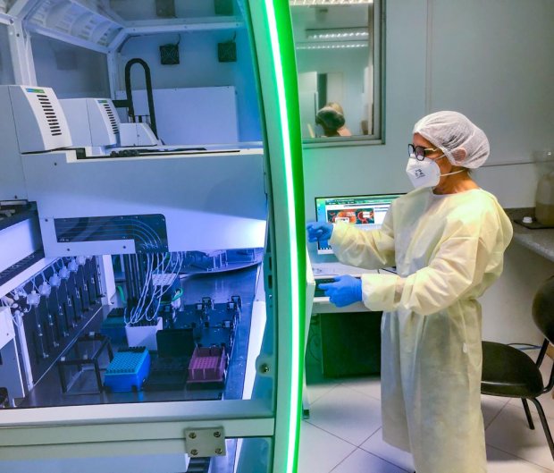 Profissional da saúde usando equipamentos de proteção individual (EPI), como máscara, touca, avental e luvas, opera uma máquina de testagem laboratorial em ambiente controlado. Ao fundo, há um computador com monitor ligado, indicando atividade no laboratório. A cena ocorre em um laboratório moderno com tecnologia avançada.