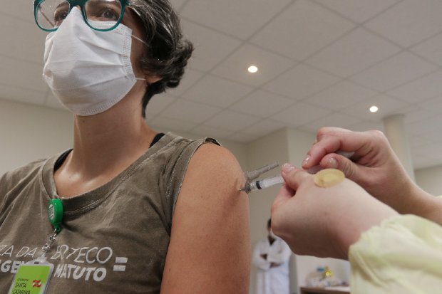 Mulher com máscara facial recebe dose de vacina contra a COVID-19 no braço esquerdo, aplicada por profissional de saúde. Ela usa camiseta com estampa e crachá do Governo de Santa Catarina. Ao fundo, há ambiente clínico com paredes claras e teto com iluminação embutida.