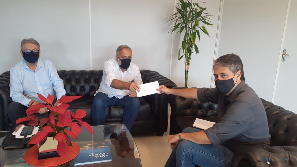 rês homens com máscaras de proteção estão sentados em sofás de couro preto, em um ambiente corporativo. Dois deles, posicionados à frente, trocam um envelope branco. 