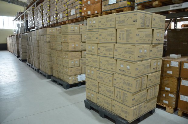 Armazém com grandes volumes de caixas de papelão empilhadas sobre paletes. As caixas estão organizadas em fileiras, indicando estoque de suprimentos, possivelmente de equipamentos médicos ou vacinas. A estrutura metálica do galpão e a iluminação natural pela janela ao fundo contribuem para a visibilidade e organização do espaço.