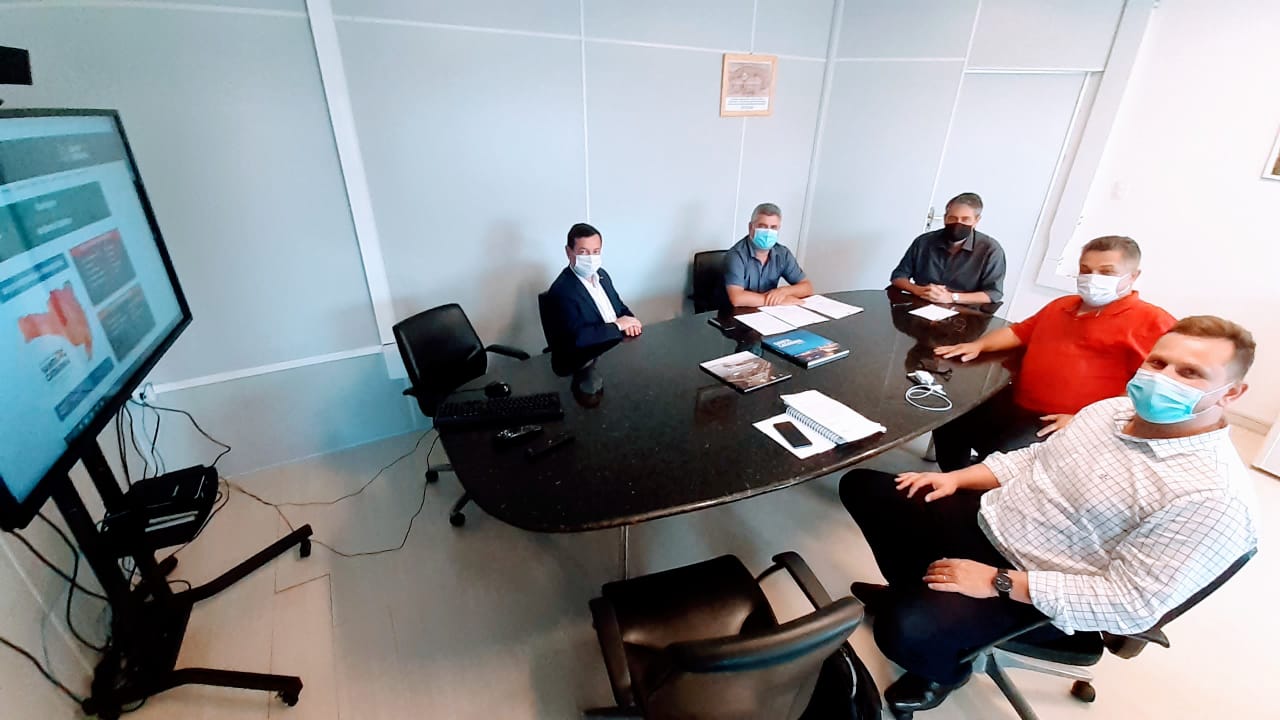 Seis homens participam de uma reunião em uma sala com mesa oval preta. Todos estão usando máscara. Há notebooks, cadernos e documentos sobre a mesa. À esquerda da imagem, um monitor exibe informações sobre a COVID-19, incluindo um mapa com cores semelhantes ao mapa de risco do estado de Santa Catarina. A reunião ocorre em um ambiente fechado, com paredes claras e boa iluminação natural.