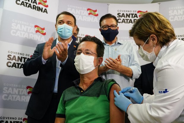 Momento da aplicação da primeira dose da vacina contra a COVID-19 em Santa Catarina. Um homem de máscara, vestido com camiseta verde, recebe a vacina no braço esquerdo aplicada por uma profissional de saúde com jaleco branco e luvas azuis. Ao fundo, autoridades estaduais aplaudem, todos usando máscaras. O cenário é decorado com painéis do Governo de Santa Catarina.