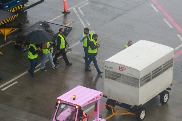 Grupo de profissionais usando coletes refletivos caminha na pista de um aeroporto em dia chuvoso. Um deles segura um guarda-chuva. À direita da imagem, um veículo reboca um compartimento branco identificado com as letras RP.