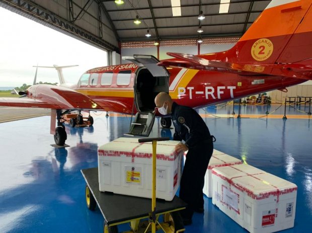 Profissional da segurança pública descarrega caixas térmicas com vacinas contra a COVID-19 de uma aeronave vermelha do Corpo de Bombeiros de Santa Catarina, identificada com o prefixo PT-RFT. A cena ocorre dentro de um hangar com piso azul brilhante, e o profissional usa máscara e uniforme escuro enquanto manuseia uma das caixas com o auxílio de um carrinho de transporte.