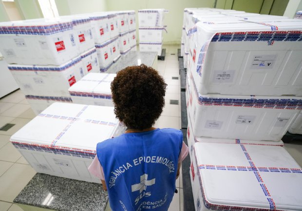 Profissional da Vigilância Epidemiológica observa caixas térmicas empilhadas com vacinas contra a COVID-19. A pessoa veste colete azul do SUS com os dizeres “Vigilância Epidemiológica – Secretaria de Vigilância em Saúde”. As caixas estão lacradas com fitas e etiquetas de transporte, organizadas em ambiente fechado com piso claro e paredes verdes.