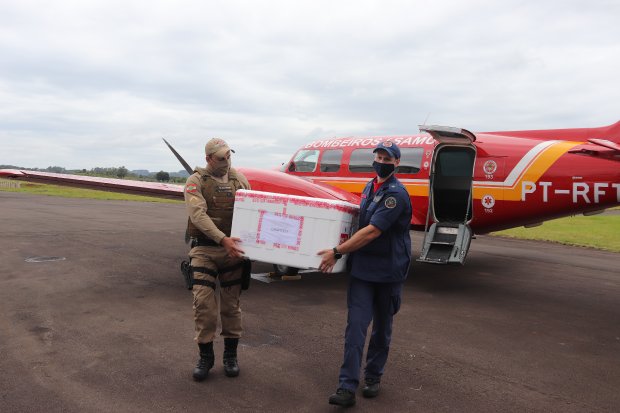 Dois profissionais uniformizados — um da Polícia Militar e outro do Corpo de Bombeiros — carregam uma caixa térmica com vacinas contra a COVID-19 em frente a uma aeronave vermelha do SAMU (PT-RFT), estacionada em uma pista de pouso. A cena mostra parte da logística de distribuição de vacinas em Santa Catarina, Brasil, durante a pandemia. O céu está nublado e o clima parece ameno.