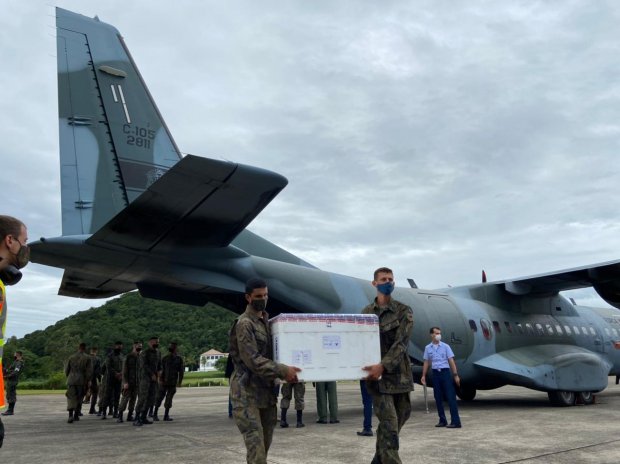 Militares das Forças Armadas descarregam uma grande caixa térmica de um avião da Força Aérea Brasileira, modelo C-105 Amazonas, em uma pista de pouso. A caixa está identificada como contendo vacinas ou insumos médicos. O cenário é de céu nublado e vegetação ao fundo, indicando uma operação logística de transporte de imunizantes.