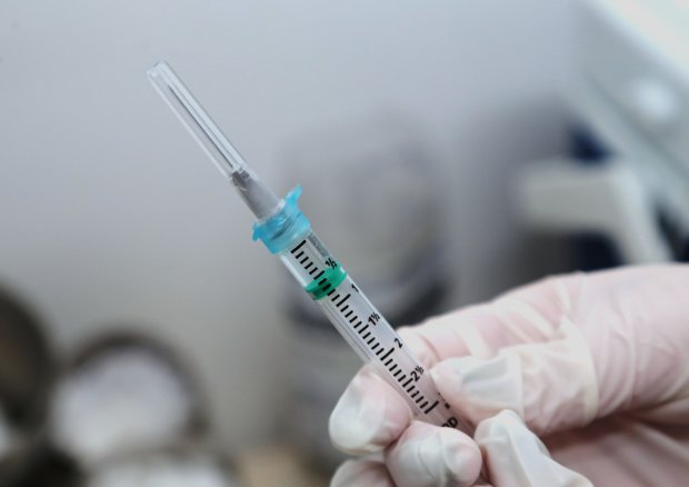 Mão com luva cirúrgica segurando uma seringa com agulha, preparada para aplicação de vacina. Ao fundo, objetos médicos desfocados, indicando ambiente clínico ou hospitalar.