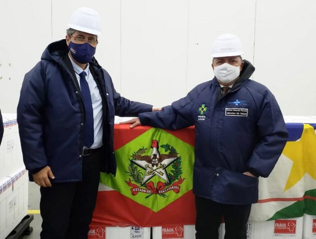 Dois homens com máscaras e capacetes brancos posam em frente a caixas térmicas utilizadas para transporte de vacinas. Ambos vestem casacos com logotipos institucionais. Ao fundo, a bandeira do estado de Santa Catarina está estendida sobre as caixas, e ao lado, parte da bandeira do estado do Mato Grosso. O ambiente sugere um centro de distribuição ou armazenamento de imunizantes.