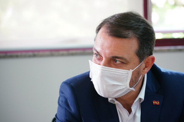 Homem de terno azul e camisa branca, usando máscara de proteção facial branca, sentado em ambiente interno com janela ao fundo. Ele possui cabelos castanhos escuros e curtos, e está com o olhar direcionado para a esquerda. Um pequeno broche com a bandeira de Santa Catarina está preso à lapela do paletó.