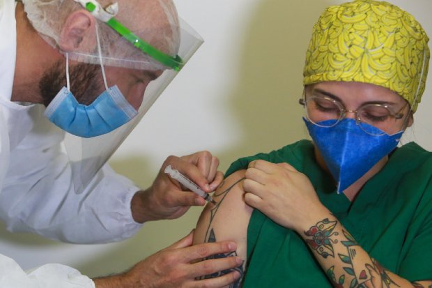 Profissional de saúde aplica uma dose da vacina contra a COVID-19 no braço de uma mulher usando jaleco verde, touca cirúrgica amarela estampada com bananas e máscara azul. A mulher tem várias tatuagens visíveis no braço. O aplicador está protegido com máscara, face shield e avental branco, demonstrando os cuidados adotados durante a imunização.