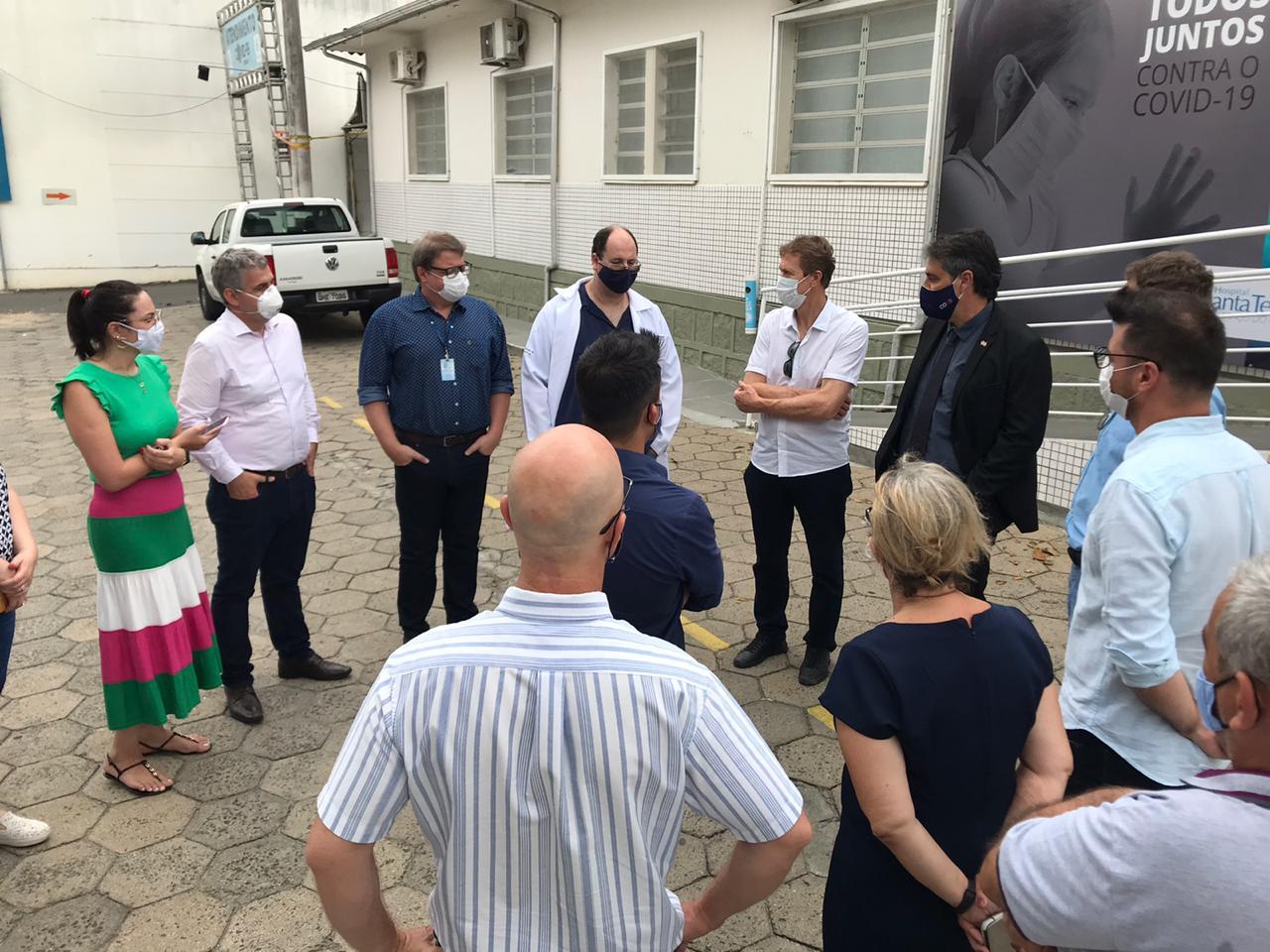 Grupo de pessoas reunidas em pé em área externa de um hospital, todas usando máscaras de proteção contra a COVID-19. Algumas usam crachás e jalecos, indicando equipe de saúde ou gestão. 