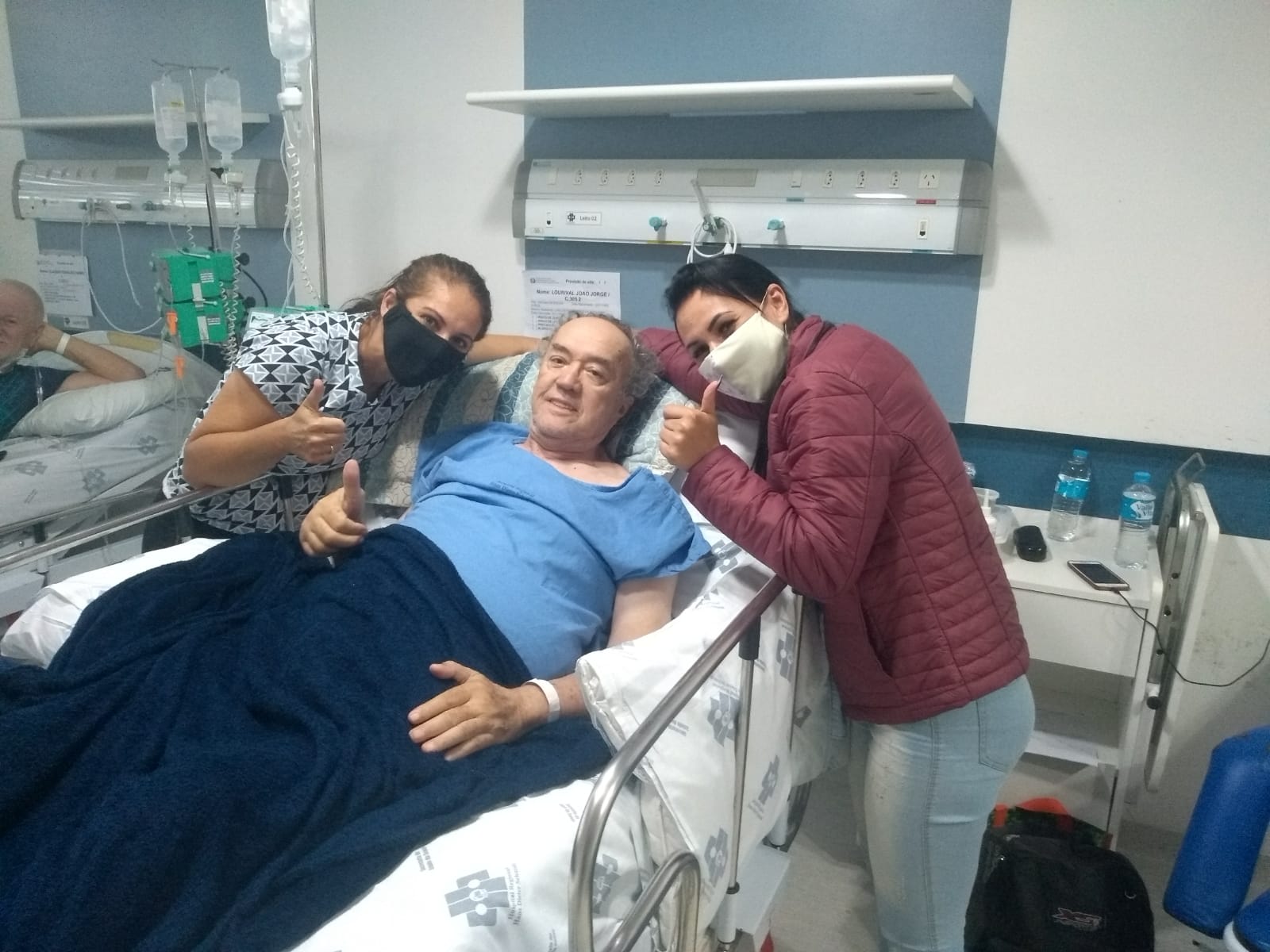 Paciente em recuperação em um leito hospitalar, sorridente e fazendo sinal de positivo com o polegar. Duas mulheres, ambas usando máscaras, estão ao lado dele, também fazendo sinal de positivo, demonstrando apoio e felicidade. Ao fundo, há outro paciente em outro leito. A cena transmite esperança e superação.