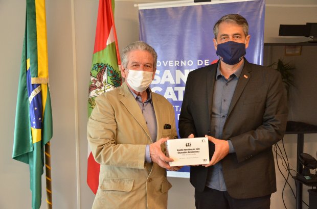 Dois homens usando máscaras faciais posam segurando uma caixa de medicamentos. Ao fundo, estão as bandeiras do Brasil e de Santa Catarina, além de um banner com o logotipo do Governo do Estado de Santa Catarina.
