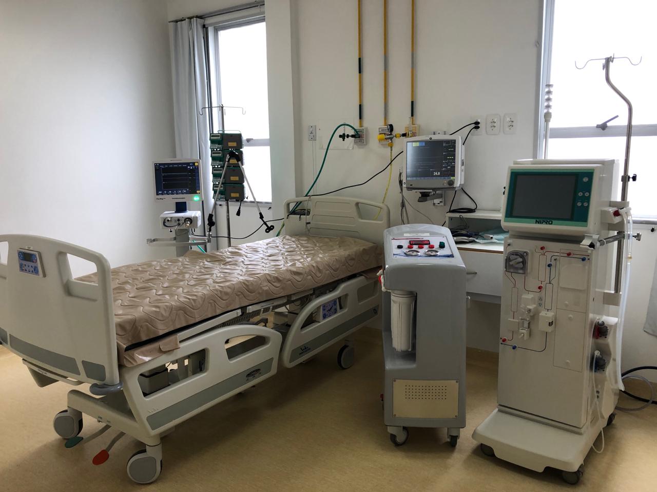  Leito hospitalar moderno com cama ajustável ao centro da imagem, cercada por equipamentos médicos como monitor cardíaco, bomba de infusão e máquina de hemodiálise. O ambiente é limpo, iluminado por luz natural vinda de duas janelas ao fundo, e possui parede branca e piso claro, típico de uma unidade de terapia intensiva (UTI).
