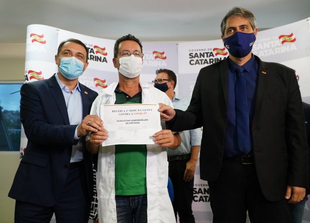 Três homens, todos usando máscaras, posam para uma foto segurando um certificado de vacinação contra a Covid-19. O homem ao centro, vestido com jaleco branco, segura o documento que indica ter recebido a primeira dose da vacina. Ao fundo, o painel exibe o logotipo do Governo do Estado de Santa Catarina, com a bandeira estadual repetida. O momento celebra simbolicamente o início da vacinação no estado.