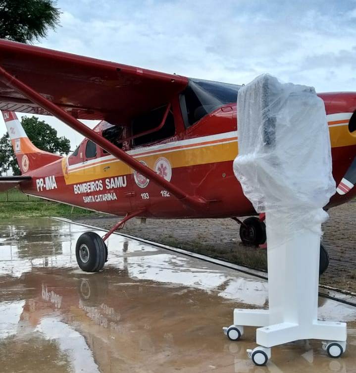 Avião vermelho do BOMBEIROS SAMU de Santa Catarina estacionado em pista molhada, com equipamento médico coberto por plástico ao lado, pronto para transporte. O céu está nublado e o solo reflete o avião devido à chuva.