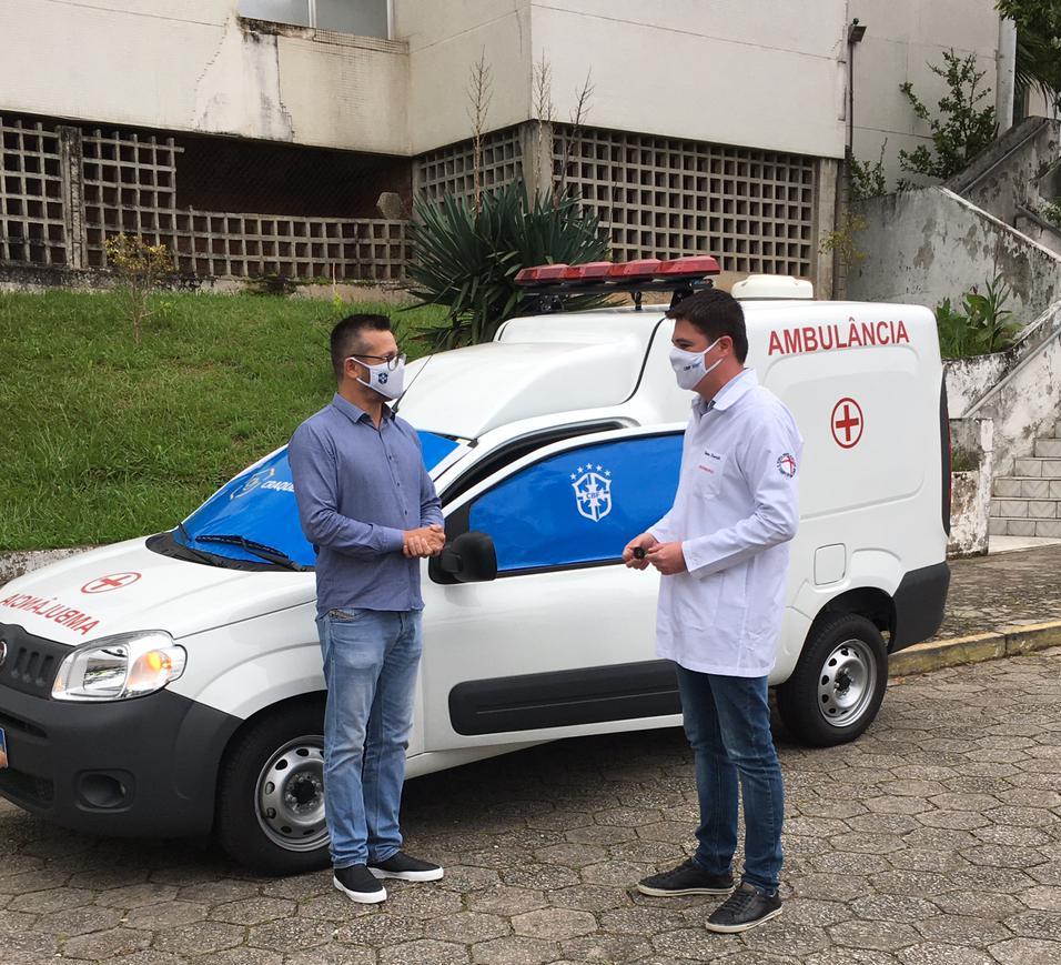 Dois representantes conversam em frente à nova ambulância doada, com janelas cobertas por tecido azul estampado com o logo da CBF, durante cerimônia de entrega em Santa Catarina.