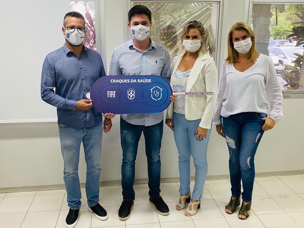 Quatro profissionais da saúde (dois homens e duas mulheres) posam com chave azul gigante escrita 'Craques da Saúde' em ambiente interno, celebrando doação de nova ambulância Fiat com apoio da CBF para transferências de pacientes.