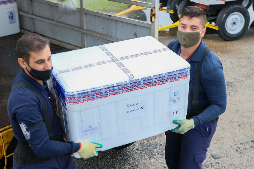 Dois funcionários vestindo macacões azuis carregando juntos uma caixa térmica com vacinas contra COVID-19, durante operação logística em Santa Catarina.