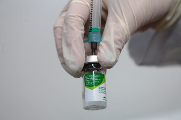 Mão enluvada de profissional de saúde segurando frasco da vacina contra gripe enquanto prepara dose com seringa.
