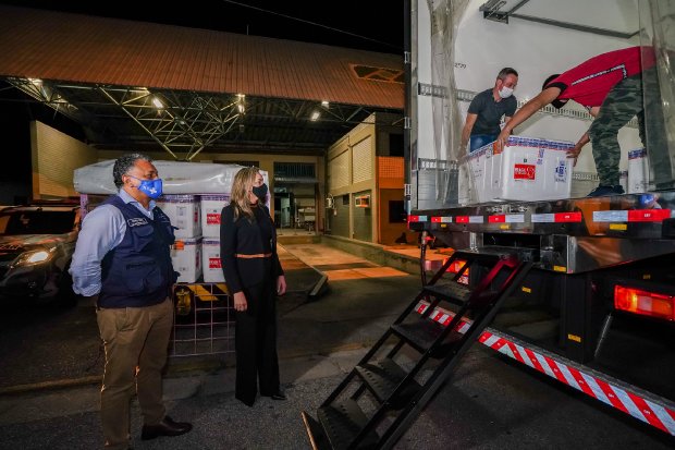 Duas pessoas observam a retirada de caixas térmicas de dentro de um caminhão refrigerado. A ação ocorre à noite, em um centro logístico. Os profissionais usam máscara e equipamentos de proteção. A cena representa a distribuição de vacinas ou insumos de saúde.