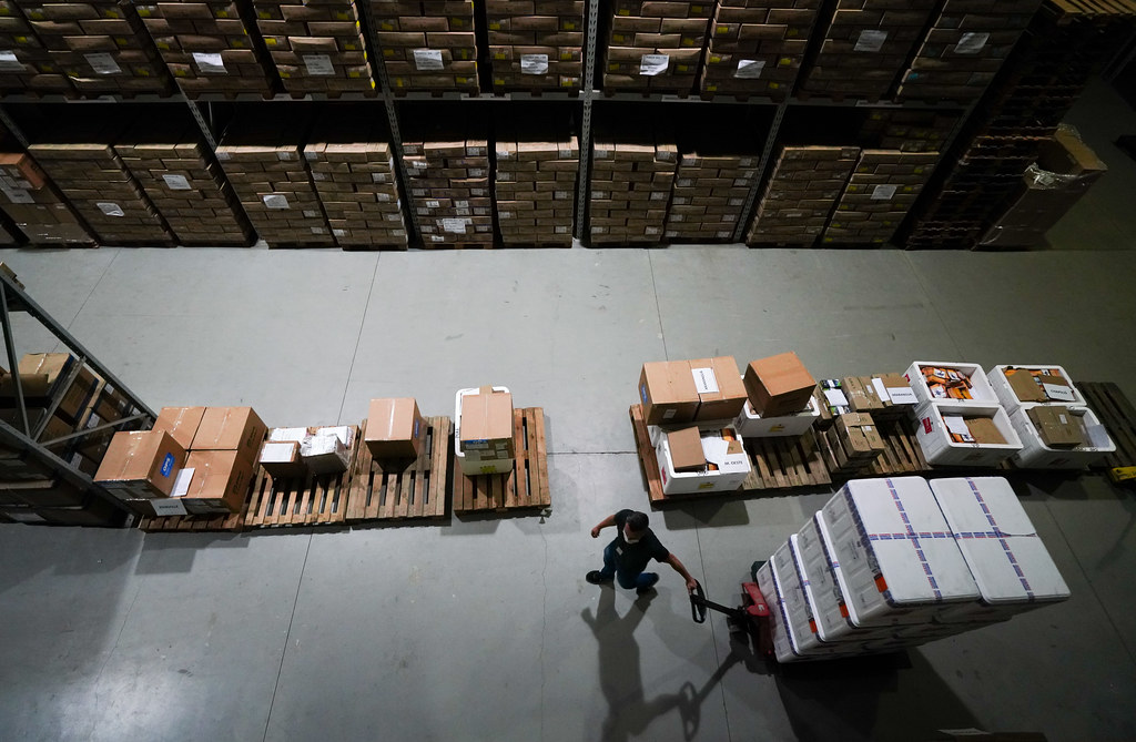 Vista aérea de um depósito com prateleiras altas repletas de caixas empilhadas. No centro, um homem movimenta caixas térmicas brancas com um carrinho manual, entre paletes organizados com diferentes volumes. A cena sugere a logística de armazenamento e distribuição de vacinas ou insumos médicos.