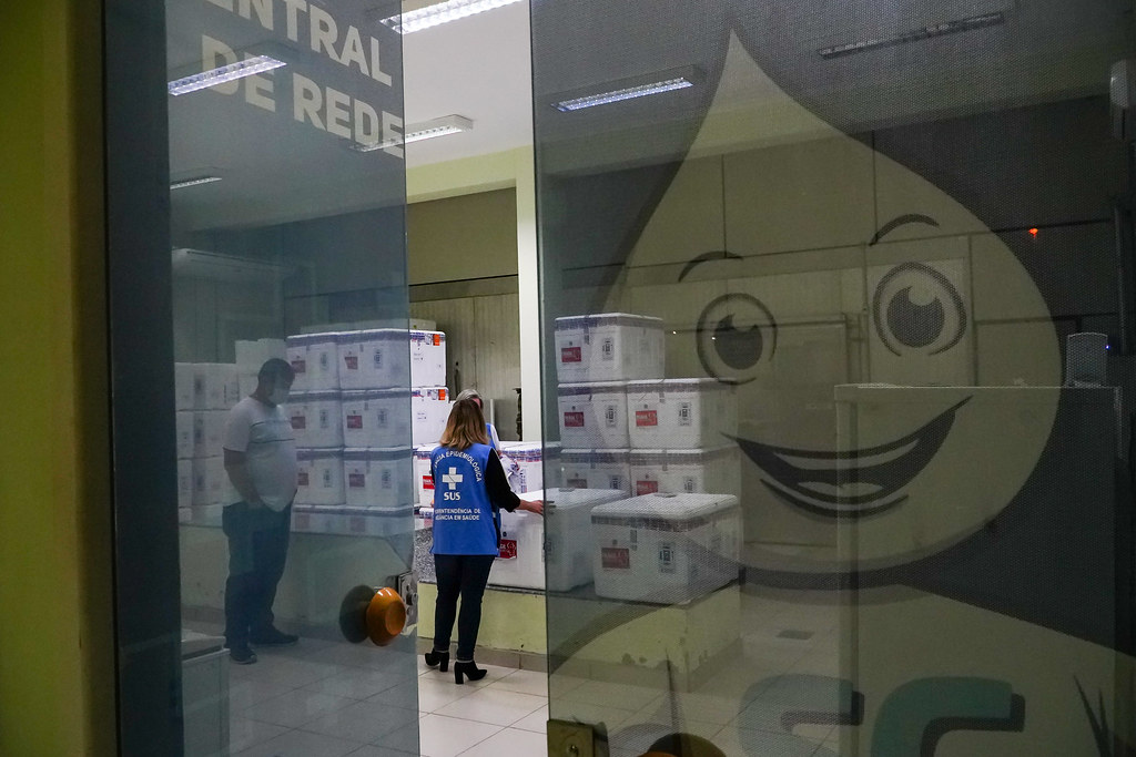 Dois profissionais da saúde conferem caixas térmicas empilhadas em uma sala com a inscrição parcial Central de Redes. A mulher usa colete do SUS com identificação da Vigilância Epidemiológica. Na porta de vidro, há a imagem reflexiva do Zé Gotinha, símbolo das campanhas de vacinação no Brasil.