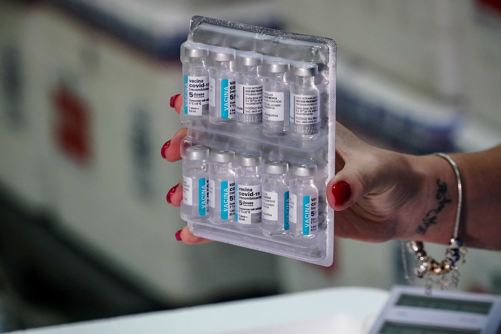 Mão de uma profissional da saúde segurando um suporte plástico com dez frascos da vacina contra a COVID-19. Os frascos têm rótulo azul e branco com a inscrição vacina covid-19 recombinante, 5 doses. Ao fundo, há caixas térmicas desfocadas utilizadas para armazenar os imunizantes.