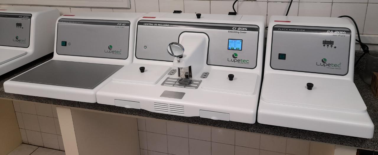 Equipamentos de laboratório da marca Lupetec organizados sobre uma bancada. Ao centro está a Central de Inclusão CI 2014, utilizada em procedimentos de histotécnica. À esquerda e à direita dela há placas aquecidas (modelos PR IQ e PA 2014), empregadas no preparo de lâminas. A parede ao fundo é revestida com azulejos brancos.