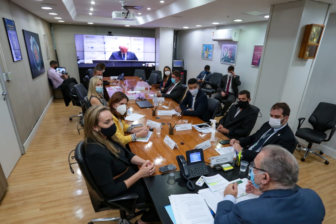 Autoridades da Secretaria da Saúde de Santa Catarina e representantes do governo reunidos em uma mesa de reunião, todos utilizando máscaras de proteção facial. A cena ocorre durante uma agenda em Brasília, em 2021, com o objetivo de discutir a habilitação de mais leitos de COVID-19 para o estado. A imagem reflete o contexto da pandemia, com medidas de segurança e esforços para ampliar a capacidade hospitalar.