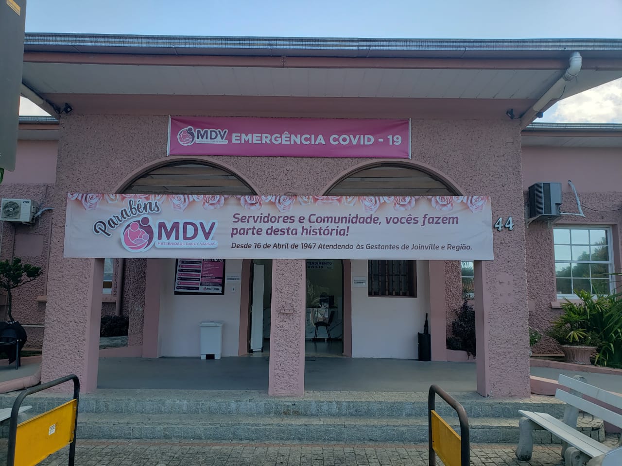 Fachada da Maternidade Darcy Vargas, um prédio hospitalar pintado na cor rosa. Na entrada, duas faixas estão instaladas: a primeira diz 'MDV Emergência COVID', destacando o setor de atendimento à pandemia; a segunda exibe a mensagem 'Parabéns MDV, servidores e comunidade - vocês fazem parte dessa história', em celebração aos profissionais e à população. A imagem retrata a união entre o combate à COVID-19 e a valorização do trabalho hospitalar durante a pandemia em 2021.