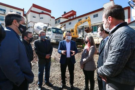 Grupo de pessoas usando máscaras conversa em um canteiro de obras, com caminhão betoneira e escavadeira ao fundo. Os prédios ao redor têm fachada clara com detalhes vermelhos. O ambiente sugere uma visita técnica ou inspeção em construção ligada à área da saúde.