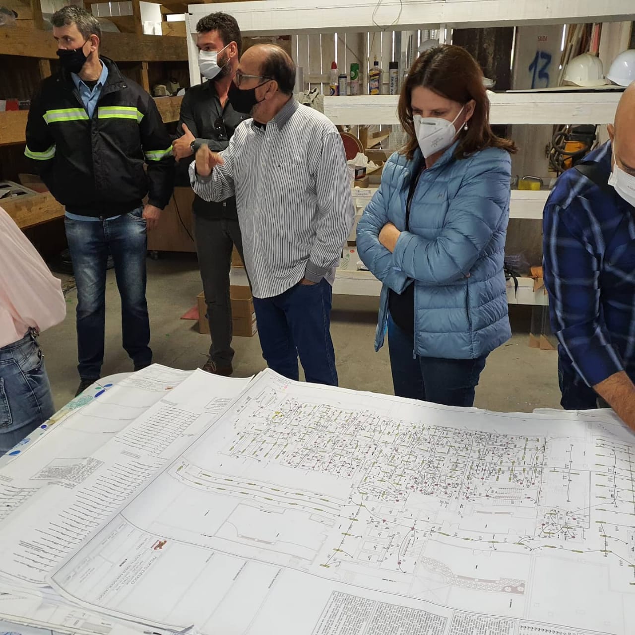 A secretária Carmen Zanotto, vestindo um casaco azul e máscara branca, analisa atentamente uma folha com desenho técnico. A cena mostra o momento em que ela avalia o projeto de instalação de um gerador de energia para a Regional de Saúde de Lages. 