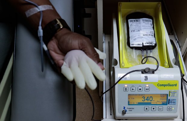 Pessoa doando sangue, com braço apoiado e conectado a um tubo que leva o sangue a uma bolsa posicionada sobre um equipamento da marca CompoGuard. A máquina exibe o peso da bolsa (340 g) e controla o processo de coleta. Uma luva de procedimento está apoiada na mão da pessoa, que utiliza um relógio de pulso.