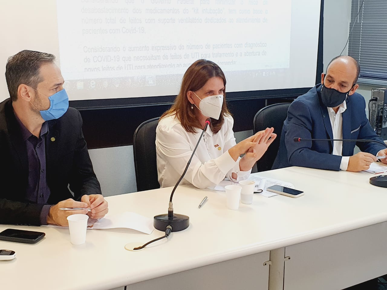 A secretária de Saúde, fala em uma mesa de reunião com dois homens, todos utilizando máscaras de proteção. Ao fundo, uma tela de projeção exibe slides.