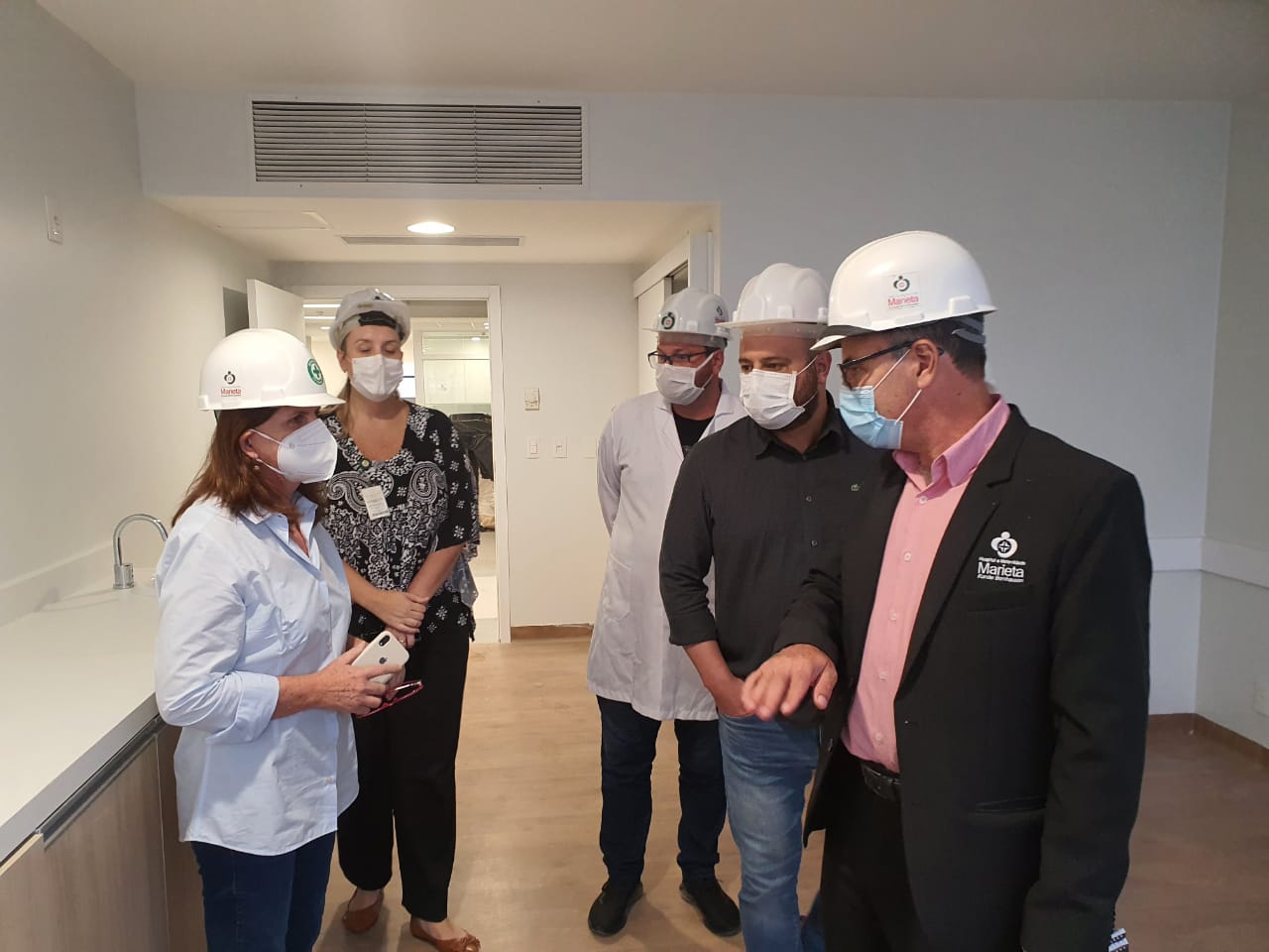 A secretária de Saúde, vestindo traje branco e máscara de proteção, conversa com quatro profissionais em um ambiente hospitalar. Todos utilizam capacetes de segurança.