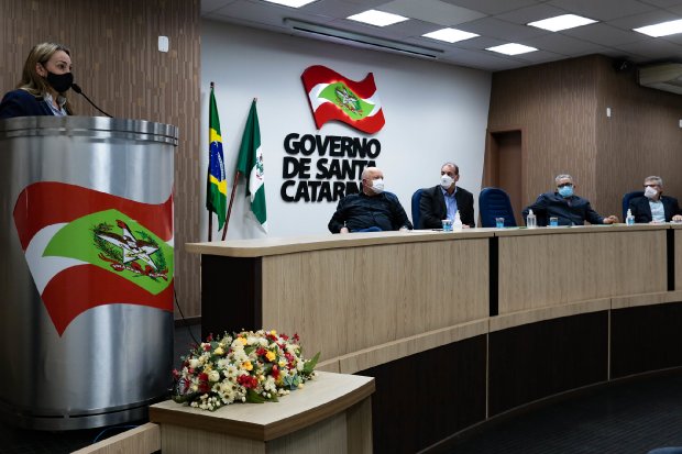 A governadora de Santa Catarina discursa em evento em Chapecó, ao lado de uma estrutura metalizada cromada com o desenho da bandeira do estado. Ela está posicionada próximo a uma mesa de madeira onde autoridades acompanham.