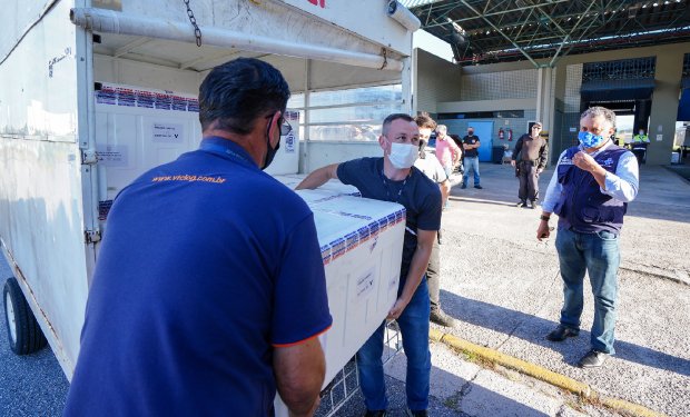 Dois profissionais vestindo uniformes azuis descarregam caixas térmicas brancas da caçamba de um caminhão, sob a supervisão de um homem de colete azul.