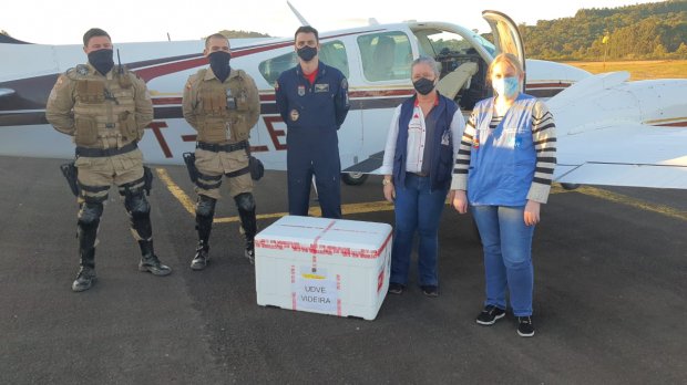 Dois militares fardados e três profissionais de saúde posam em frente a um avião, com uma caixa térmica branca no chão à frente do grupo.