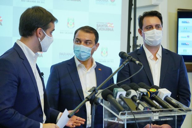 Três governadores da região Sul (PR, SC e RS) em coletiva de imprensa conjunta, com diversos microfones de emissoras na frente, anunciando medidas cooperativas no enfrentamento à pandemia.