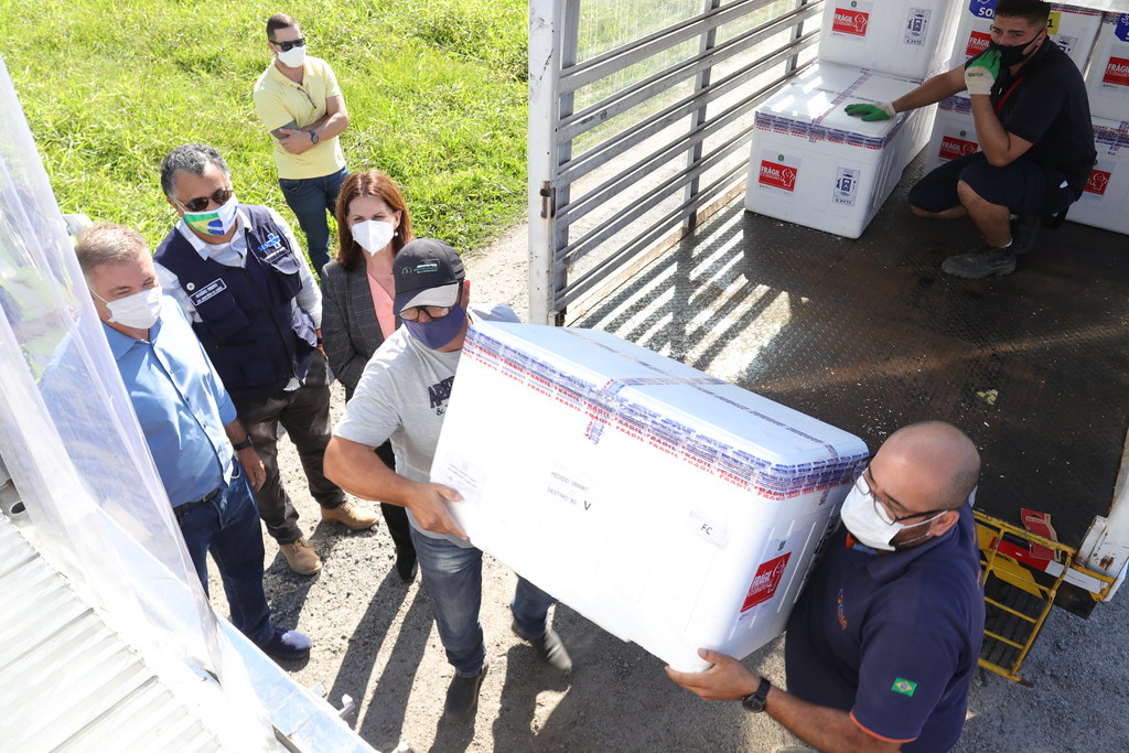 Duas pessoas descarregam caixas térmicas brancas da caçamba de um caminhão, enquanto três acompanham a operação – entre elas, Gean Loureiro, prefeito de Florianópolis, e a secretária municipal da Saúde.