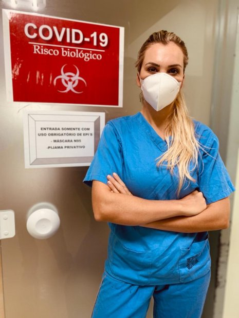 Enfermeira Luciane Buchner usando máscara de proteção em frente à porta com placa de alerta 'COVID-19 - Risco Biológico'.