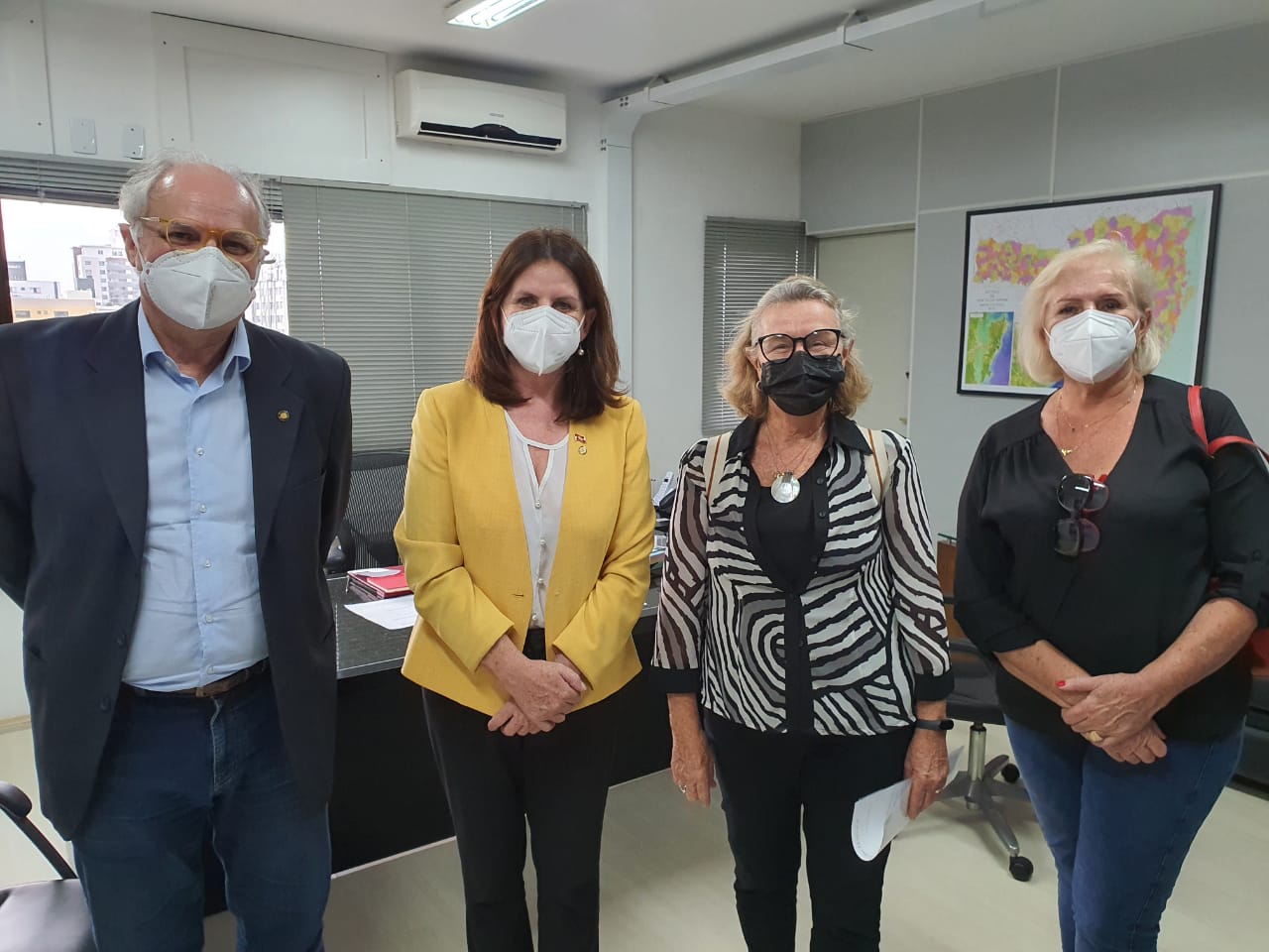 A secretária da Saúde de Santa Catarina, vestindo um blazer amarelo, posa em ambiente interno com outras três pessoas, durante reunião com representantes da FAPAES (Federação das APAEs).