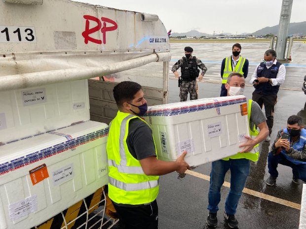 Duas pessoas vestindo coletes amarelos de identificação carregam caixas térmicas brancas, contendo 138,9 mil doses da vacina AstraZeneca/Fiocruz, em uma operação de recebimento no Governo do Estado de Santa Catarina (2021).