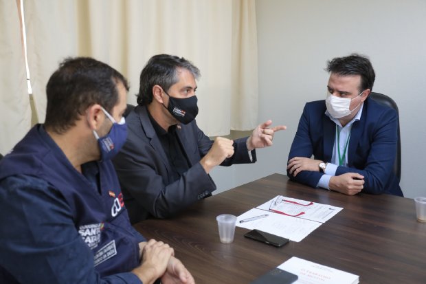 Três homens sentados em uma mesa durante reunião, todos usando máscaras; um deles gesticula enquanto fala. Sobre a mesa há papéis, copos plásticos e um celular.