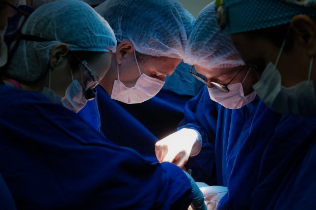 Quatro médicos em uma sala de cirurgia com iluminação focada, observando atentamente um procedimento cirúrgico. Apenas seus rostos são visíveis, iluminados pela luz fria do centro cirúrgico, enquanto o ambiente ao redor permanece escuro.